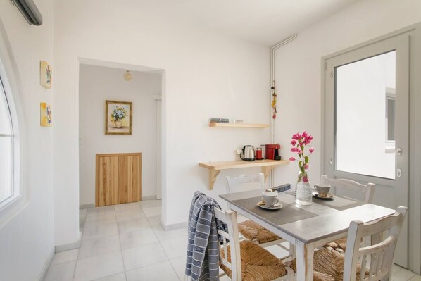 A bright, modern kitchen with white cabinetry, a wooden dining table, and a view of the city through a large window. - Maisonette whith amazing sea view. (Paros)