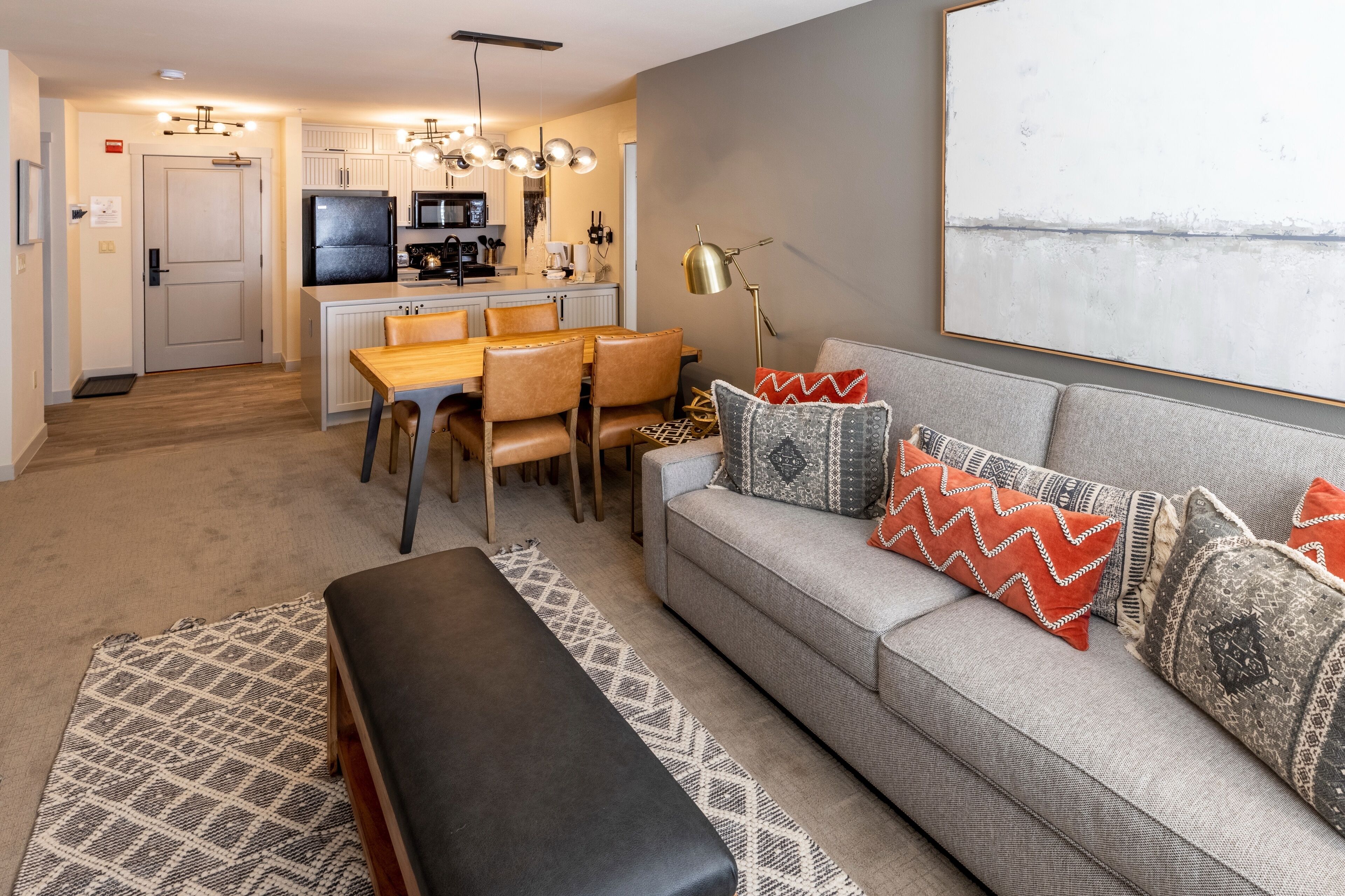 A modern living room with a grey sofa, patterned rug, and a bench. A kitchen with white cabinets and a dining area with a wooden table and chairs is visible in the background.