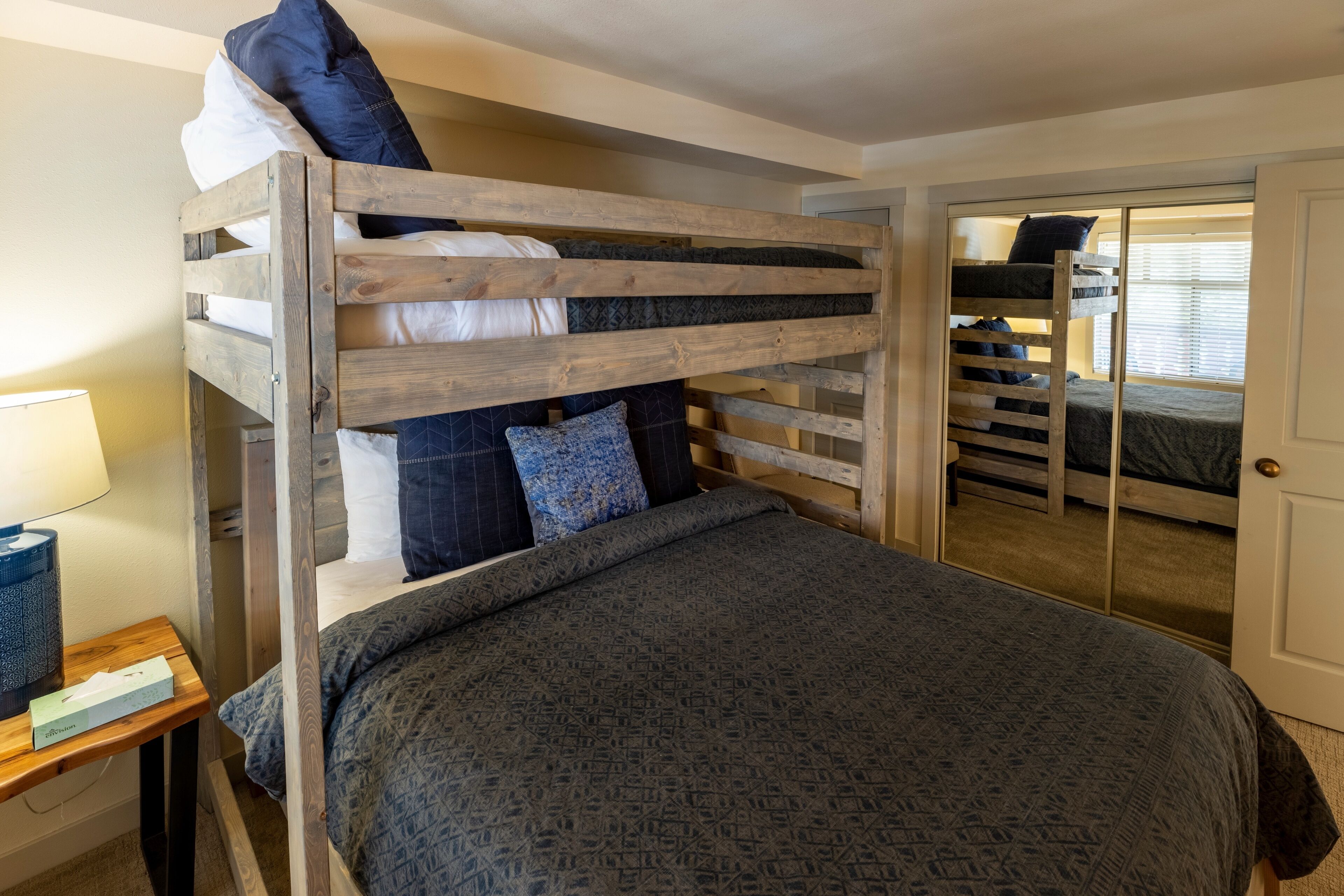 Une chambre avec un lit superposé en bois, un lit inférieur avec une couvre-lit à motifs, une table de chevet avec une lampe et un miroir.