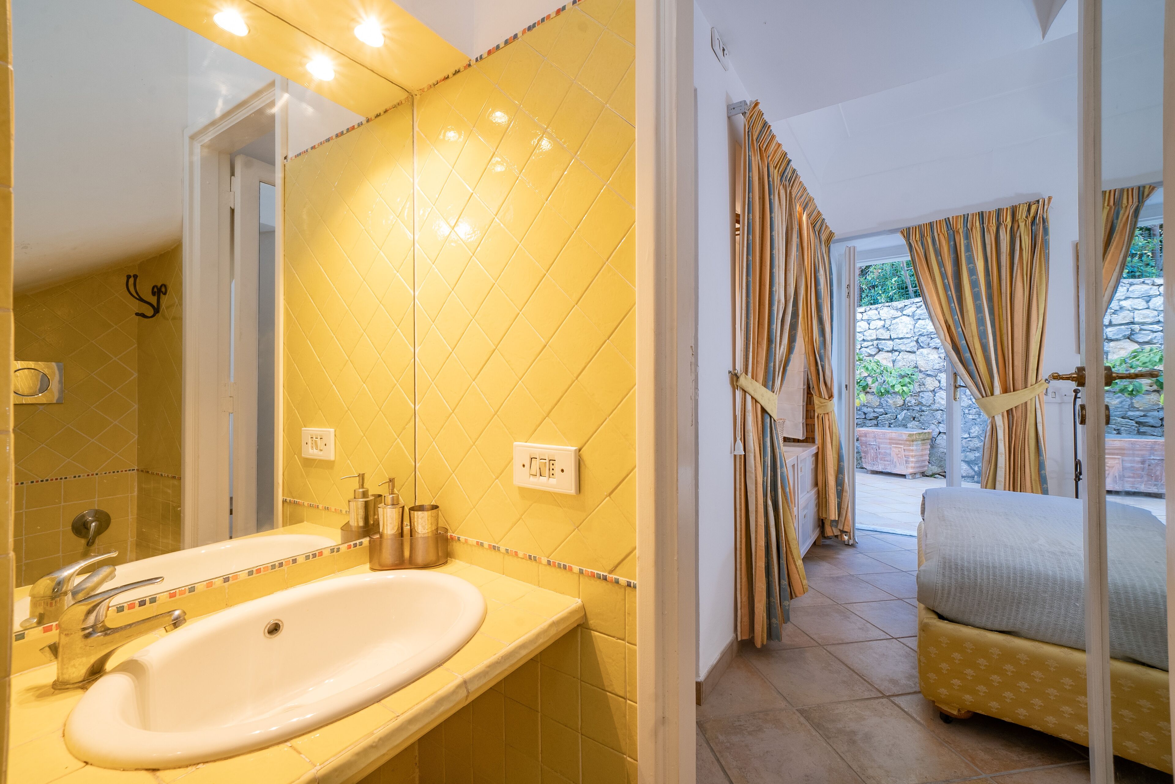 Une salle de bain avec un mur recouvert de carreaux jaunes, un lavabo blanc et un miroir. Dans la pièce adjacente, on trouve un lit avec une couvre-lit jaune et des rideaux.