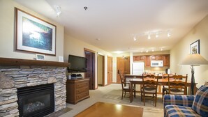 A living room with a stone fireplace, a television, a dining area with wooden chairs, and a kitchen in the background. - 3212 - One Bedroom Standard Powderhorn Lodge 1 Condo by RedAwning (Salt Lake City)