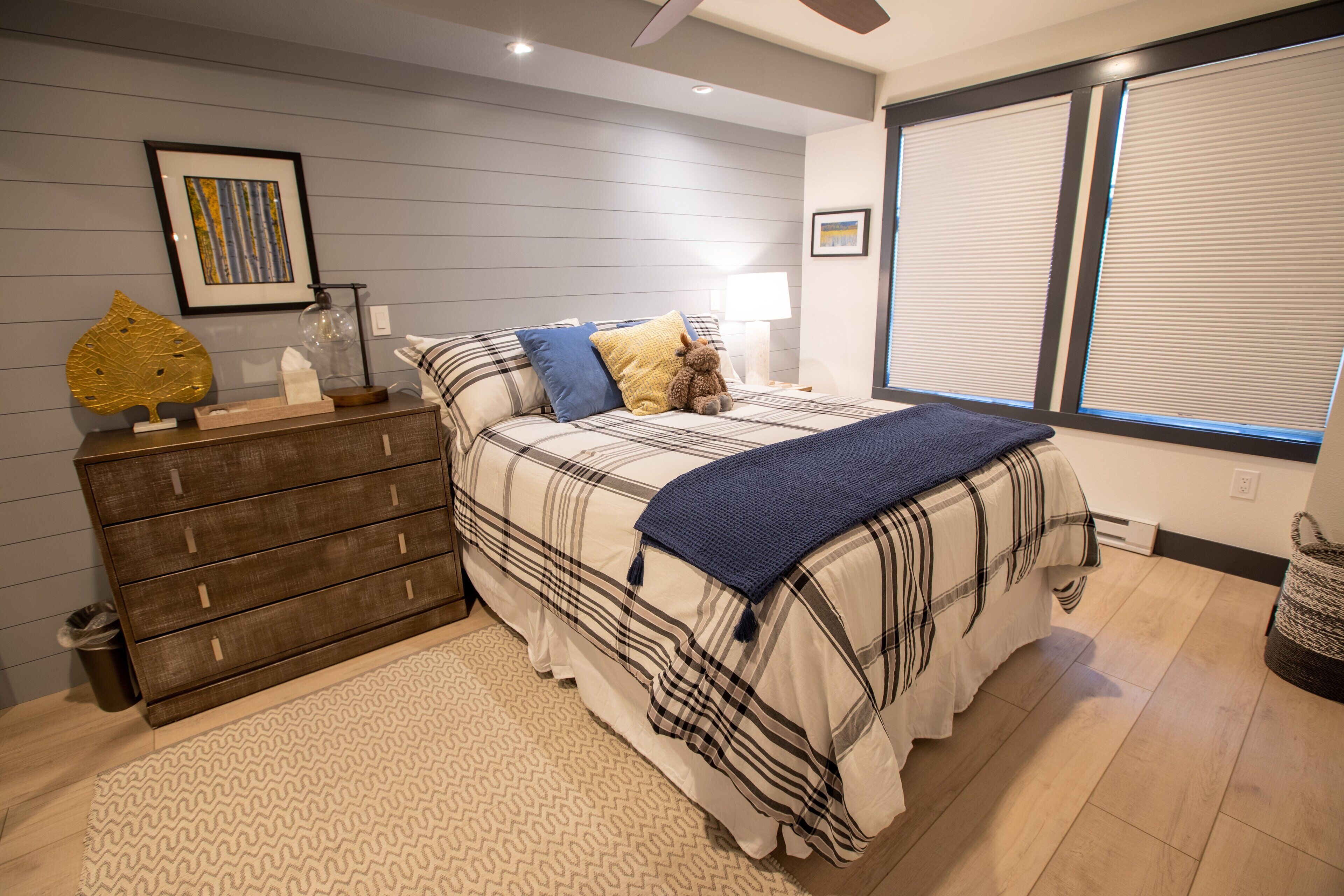 A bedroom with a bed, dresser, bedside table, lamp, and window with blinds.