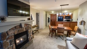 A living room with a stone fireplace, a flat-screen TV mounted above it, and a dining area with a wooden table and chairs. - 2214 - Two Bedroom Standard Eagle Springs East 2 Condo by RedAwning (Solitude)