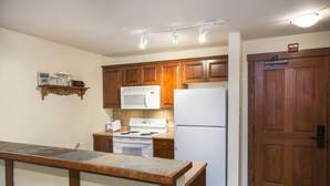 Une cuisine avec des armoires en bois, un réfrigérateur blanc, une cuisinière et un four à micro-ondes. Il y a une étagère en bois au-dessus de la cuisinière et une porte sur le côté droit.