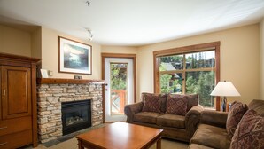 A living room with a stone fireplace, a wooden coffee table, a brown leather sofa, and a large window with a view of trees. - 3316 - One Bedroom + Den Standard Powderhorn Lodge 1 Condo by RedAwning (Salt Lake City)