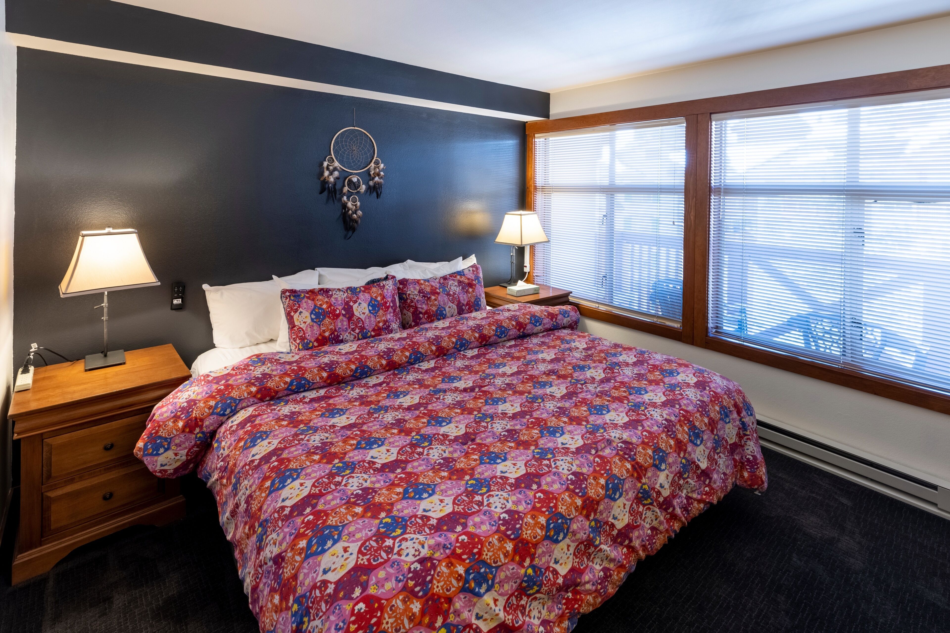 A bedroom with a large bed, two bedside tables, a dreamcatcher on the wall, and a window with blinds.