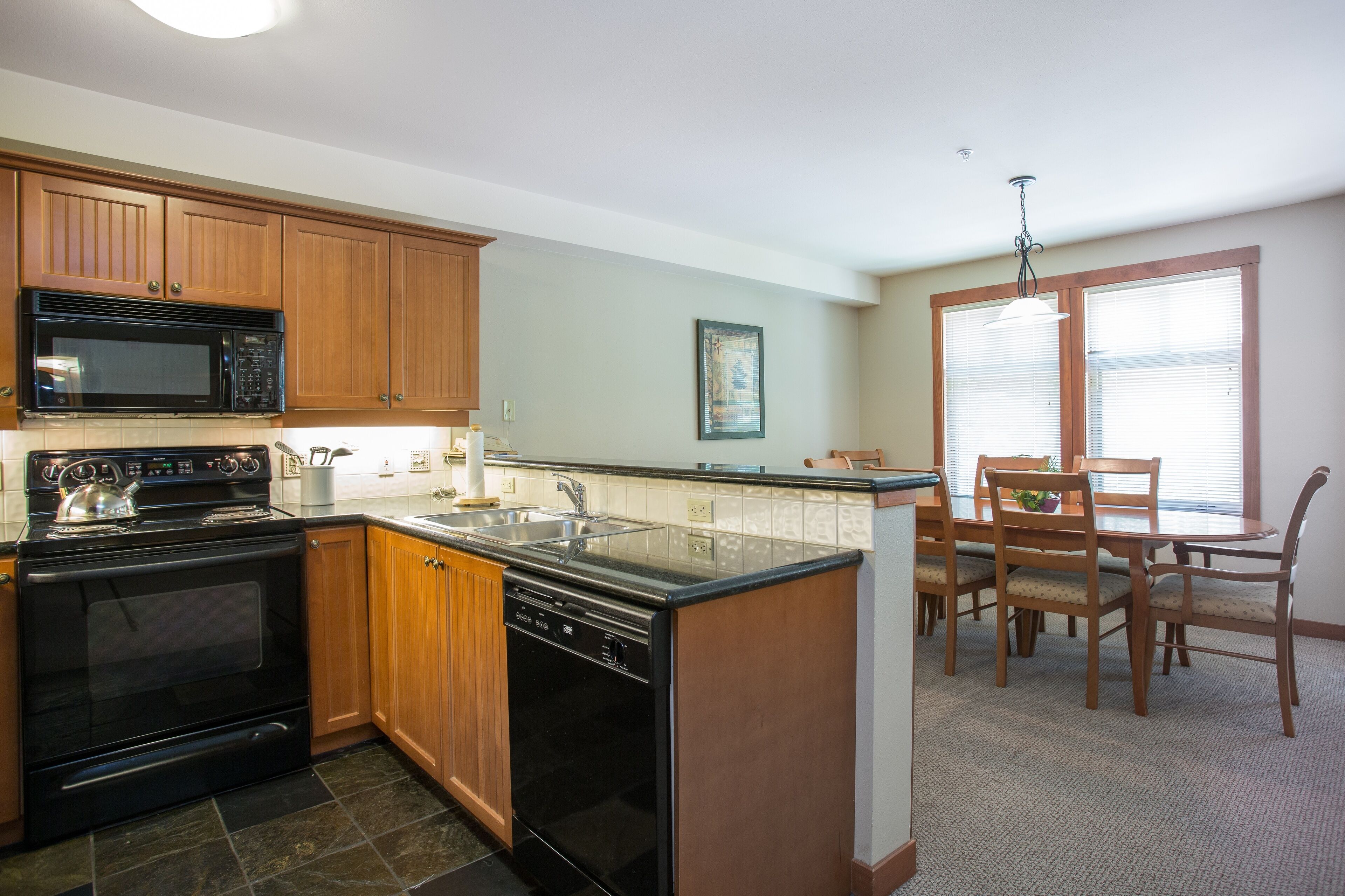 A kitchen with wooden cabinets, a black microwave, a stove, and a dishwasher. There is a dining area with a table and chairs.