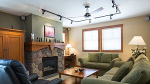 Un salon avec une cheminée, un canapé en cuir, un canapé vert, une table basse en bois et un ventilateur de plafond.