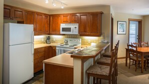 A kitchen with wooden cabinets, a white refrigerator, a microwave, a stove, and a dining area with a table and chairs. - 3219 - Three Bedroom Standard Powderhorn Lodge 3 Condo by RedAwning (Salt Lake City)
