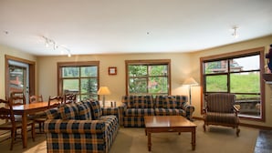 A living room with a sofa, armchair, coffee table, and dining set. There are large windows with views of greenery and a building. - 3219 - Three Bedroom Standard Powderhorn Lodge 3 Condo by RedAwning (Salt Lake City)