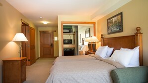 Habitación de hotel con una cama grande, dos mesitas de noche con lámparas, un cabecero de madera, un cuadro en la pared y vistas al área de la cocina.
