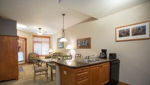 A kitchen with wooden cabinets, a sink, and a coffee maker. There is a dining area with a table and chairs, and a window with blinds.