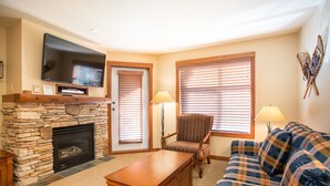 A living room with a stone fireplace, a flat-screen TV mounted above it, a plaid sofa, a striped armchair, and two lamps. - 3216 - One Bedroom + Den Standard Powderhorn Lodge 1 Condo by RedAwning (Salt Lake City)