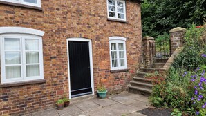 A brick house with a black door, white-framed windows, and a small garden with potted plants and flowers. - Lovely 2-bed Cottage in Coalbrookdale Telford (Telford)