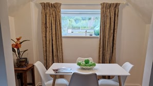 A dining area with a white table, four chairs, a bowl of green apples, a vase with flowers, and a window with curtains. - Lovely 2-bed Cottage in Coalbrookdale Telford (Telford)