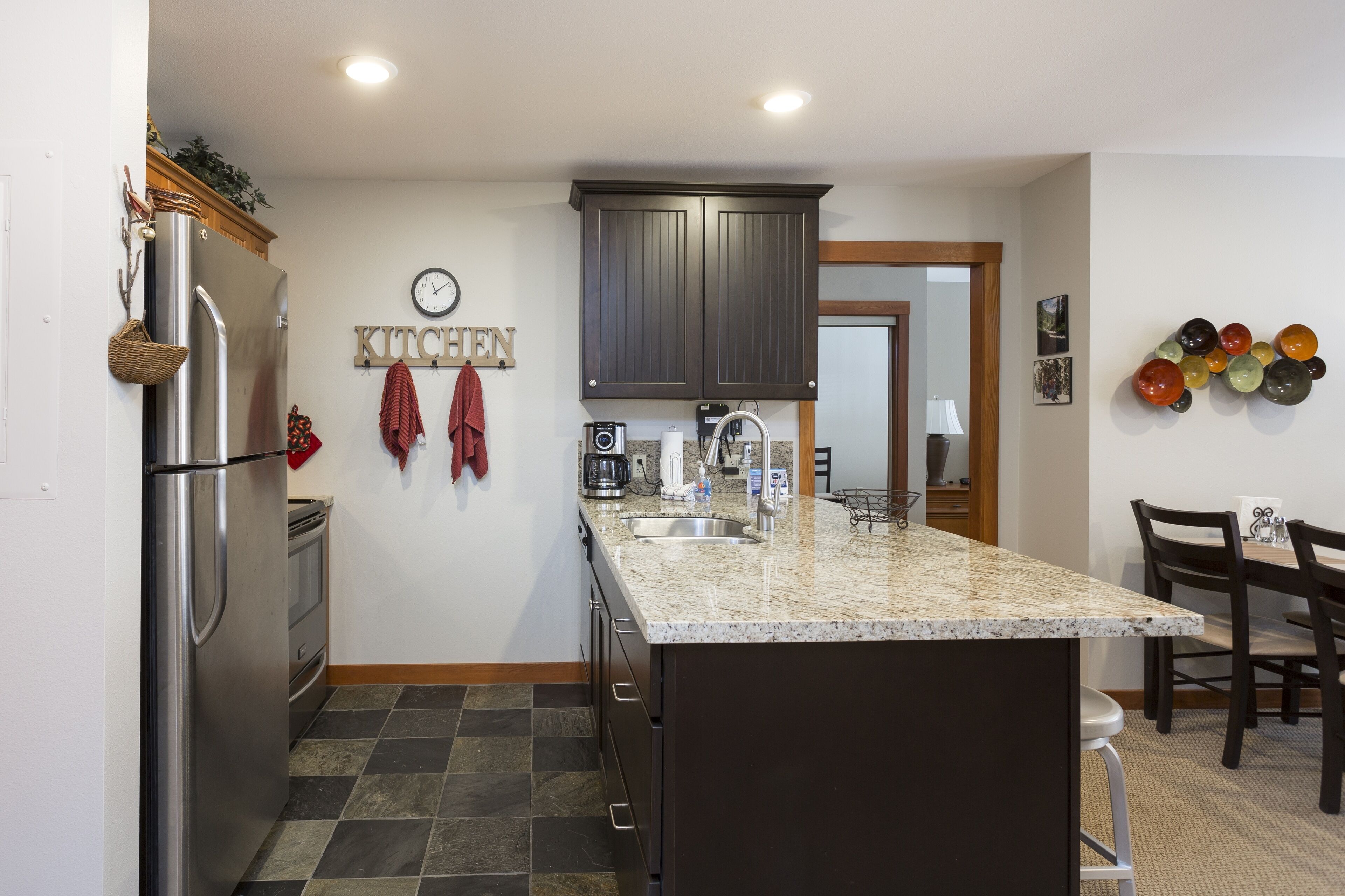 A kitchen with a granite countertop, stainless steel refrigerator, dark wooden cabinets, a clock, and a wall-mounted art piece.