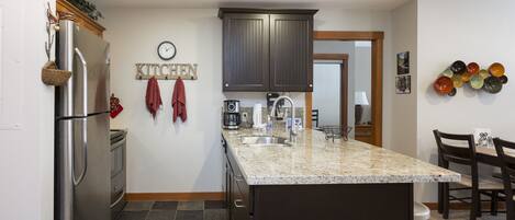 A kitchen with a granite countertop, stainless steel refrigerator, dark wooden cabinets, a clock, and a wall-mounted art piece.