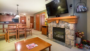 A living room with a stone fireplace, a flat-screen TV mounted above it, wooden furniture, and a kitchen area in the background.