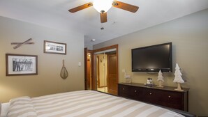 Quarto com cama, ventoinha no teto, TV de tela plana, cômoda e quadros na parede.