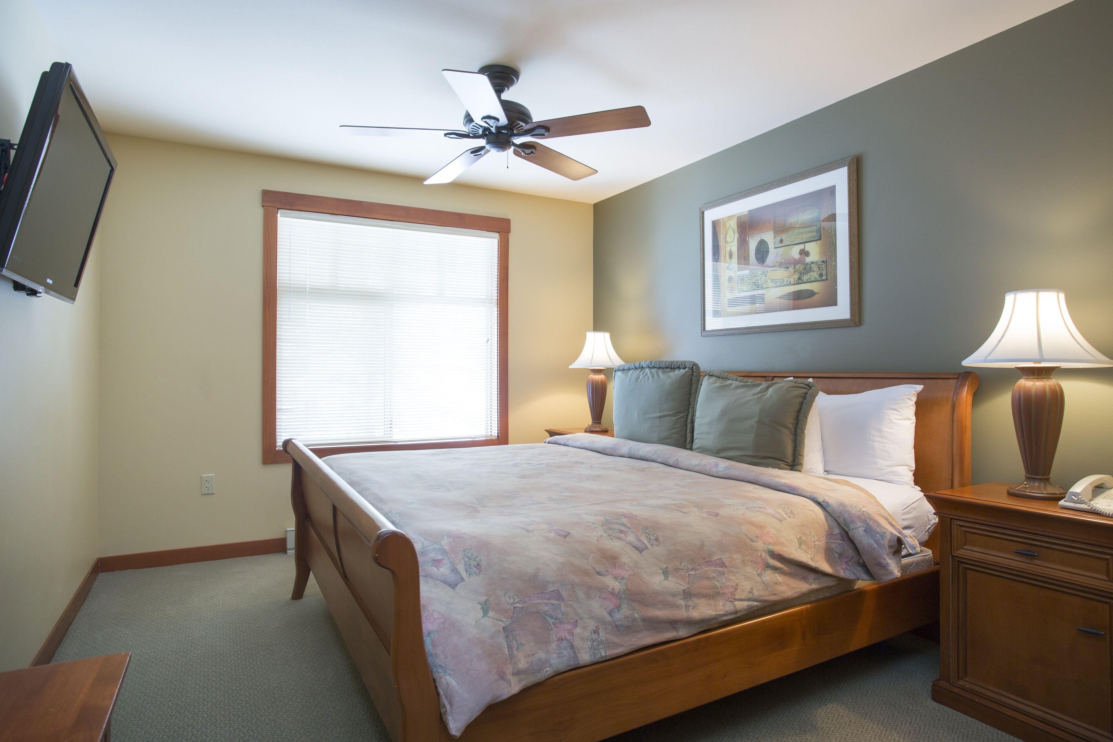 A bedroom with a bed, bedside lamps, a ceiling fan, a window, and a wall-mounted TV.