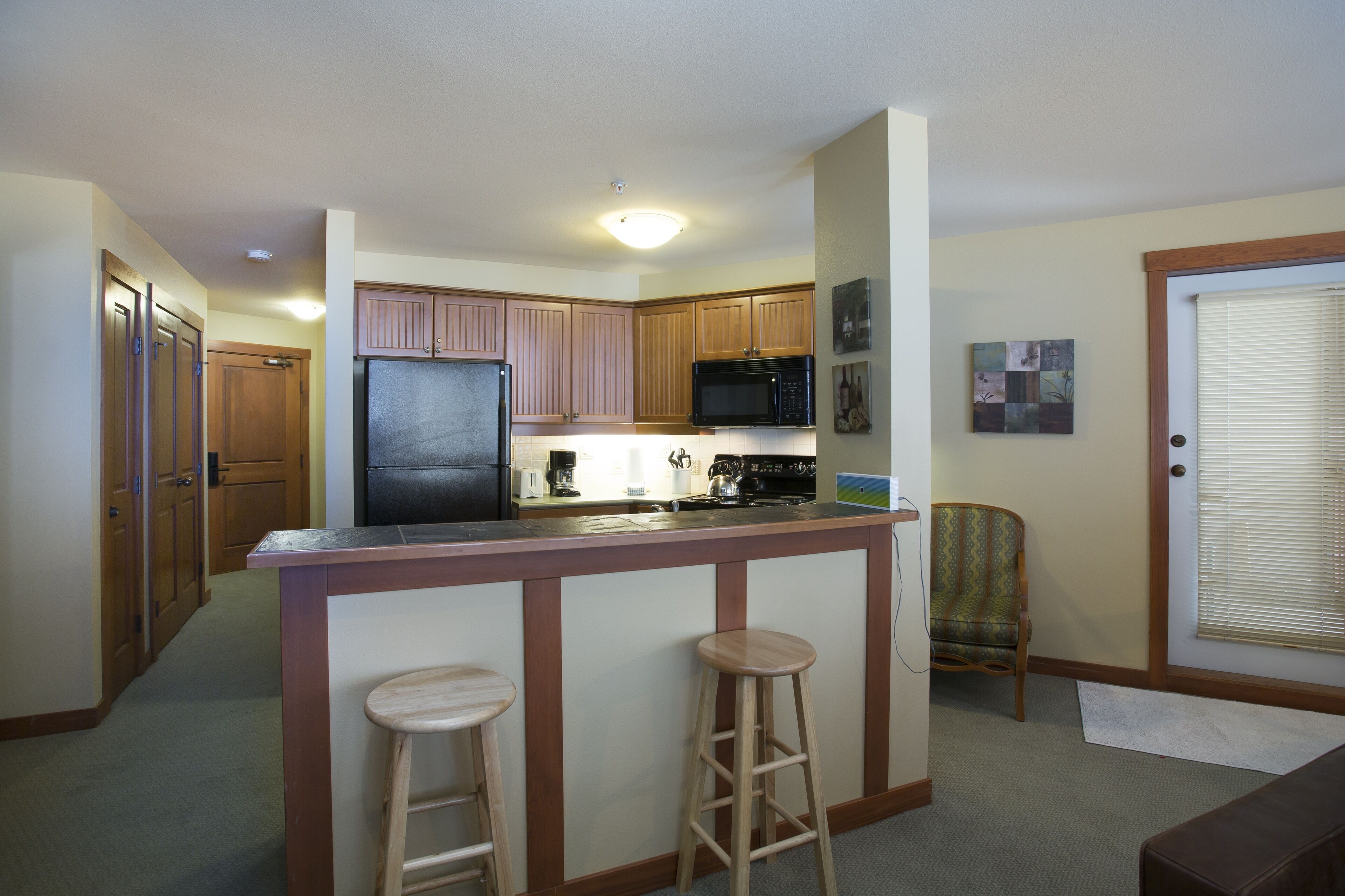 A compact kitchen with wooden cabinets, a refrigerator, a microwave, and a bar stool.