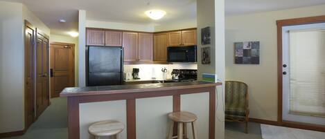 A compact kitchen with wooden cabinets, a refrigerator, a microwave, and a bar stool.