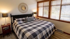 Une chambre à coucher avec une couvre-lit à carreaux, deux lampes de chevet, un miroir rond accroché au mur et une fenêtre avec des stores.