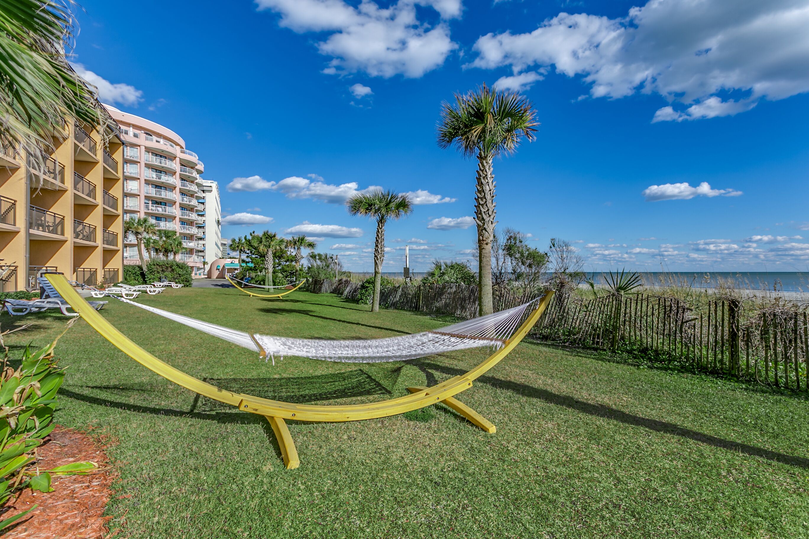 A hammock on a grassy area with a view of a beach and a hotel building in the background.