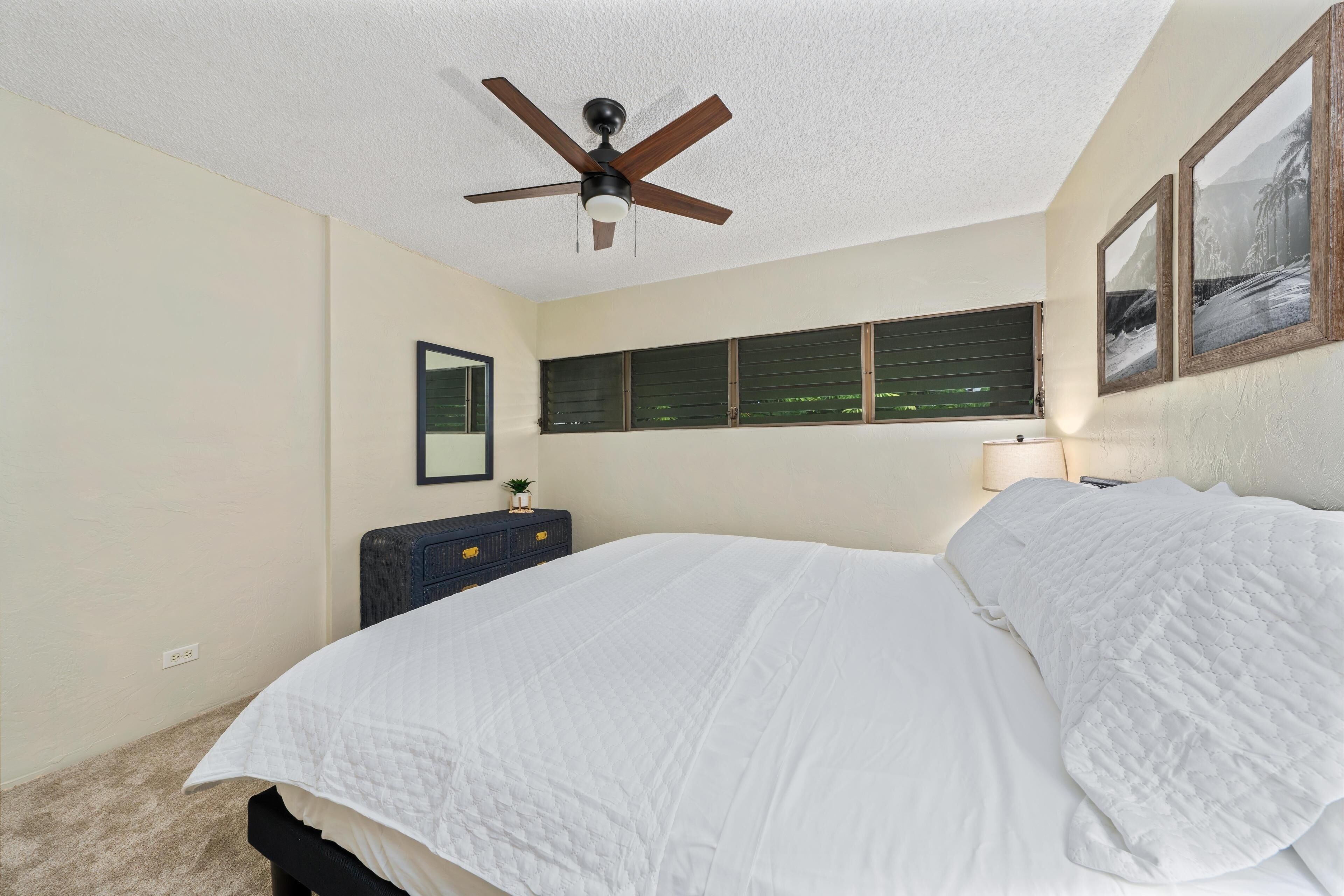 A bedroom with a bed, ceiling fan, window with blinds, dresser, and wall-mounted lamp.