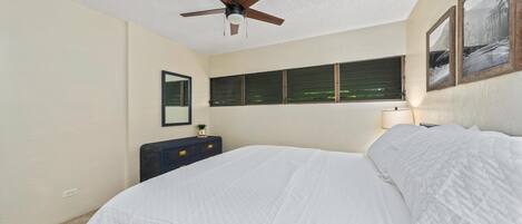 A bedroom with a bed, ceiling fan, window with blinds, dresser, and wall-mounted lamp.