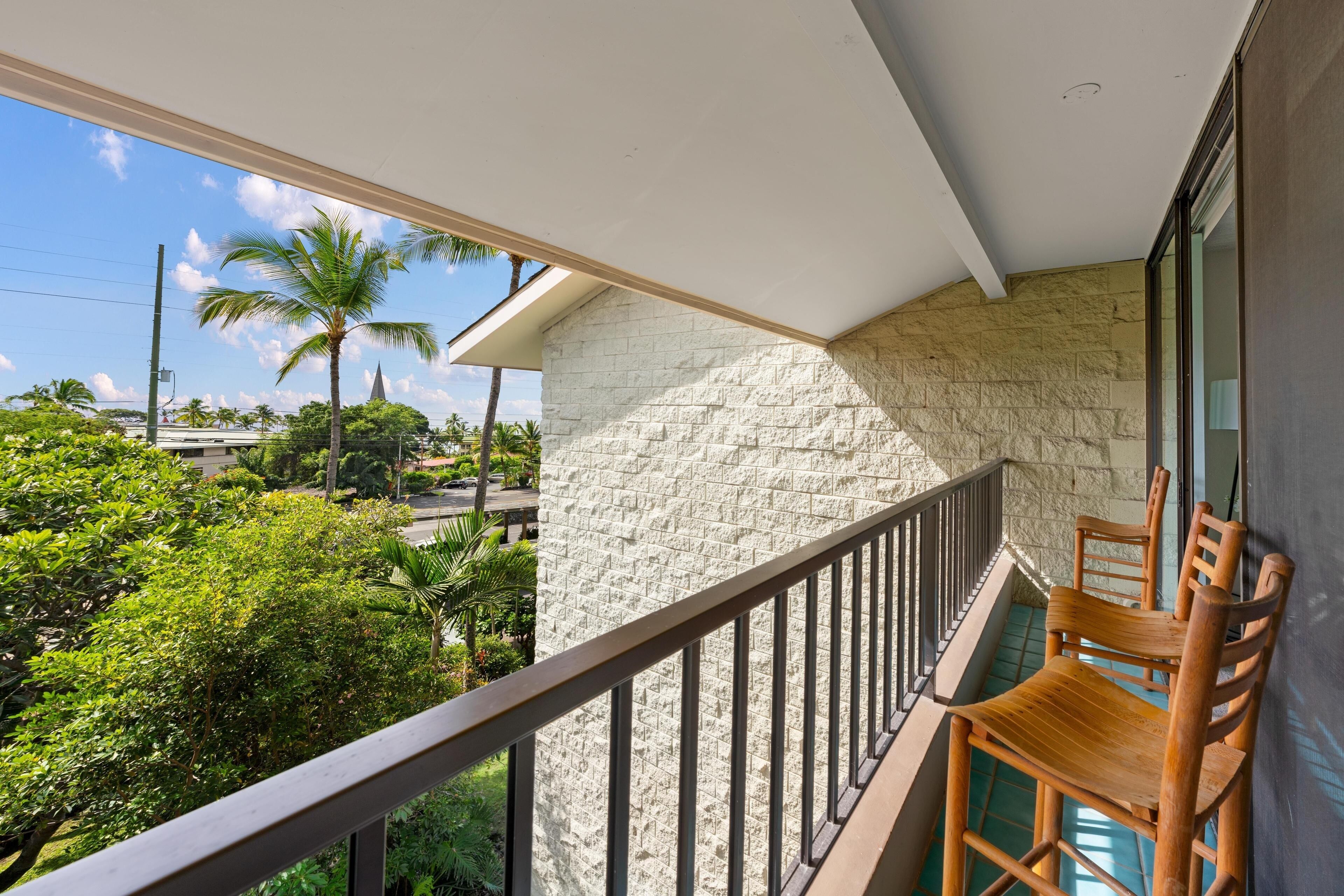 A balcony with a stone wall, wooden chairs, and a view of greenery and buildings.