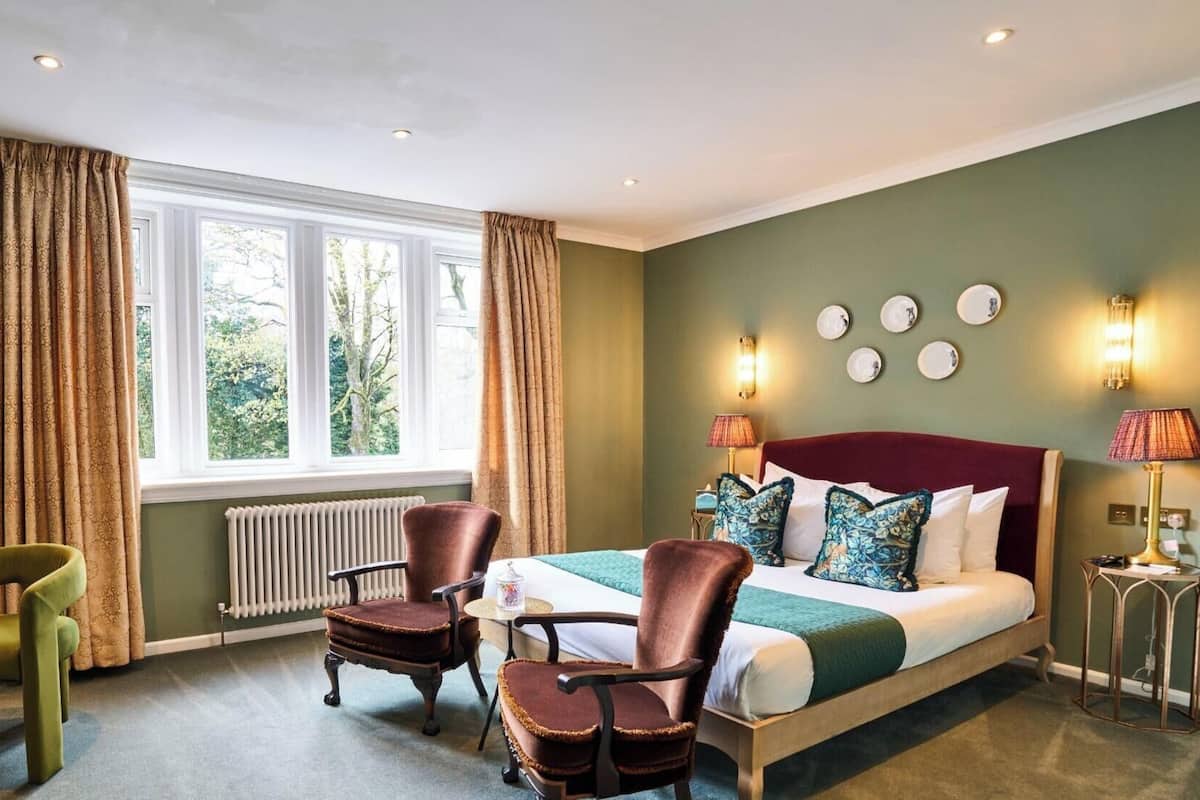 Quarto de hotel com cama, duas cadeiras, mesa, janela com cortinas, radiador e luminárias fixadas na parede.