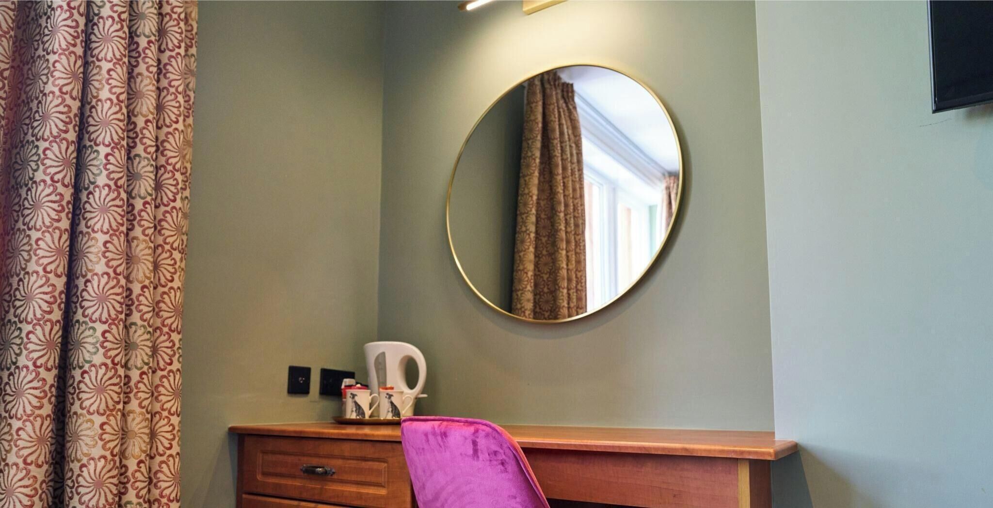 Una habitación con un aparador de madera, un espejo, una silla morada y cortinas estampadas.