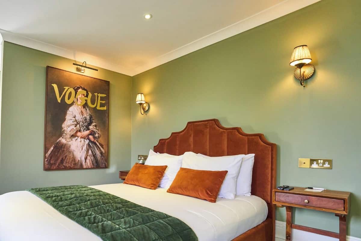 A hotel room with a large bed, a bedside table, a nightstand, a poster on the wall, and a window.