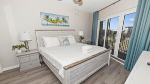 A bedroom with a wooden bed, white bedding, a nightstand with a lamp, a painting of palm trees, and a balcony with blue curtains.