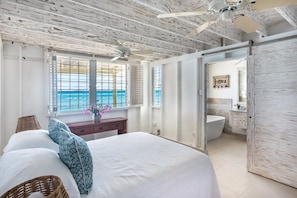 A bedroom with a large bed, a ceiling fan, a view of the ocean through the window, and a bathroom with a bathtub. - Sunset Reach by Blue Sky Luxury (Mullins)