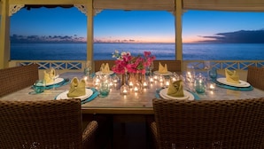 A dining table set for four with wicker chairs, a vase of flowers, and candles. - Easy Reach by Blue Sky Luxury (Mullins)