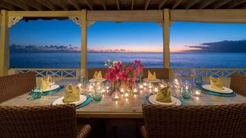 A dining area with wicker chairs, a table set for two, and a view of the ocean at dusk.