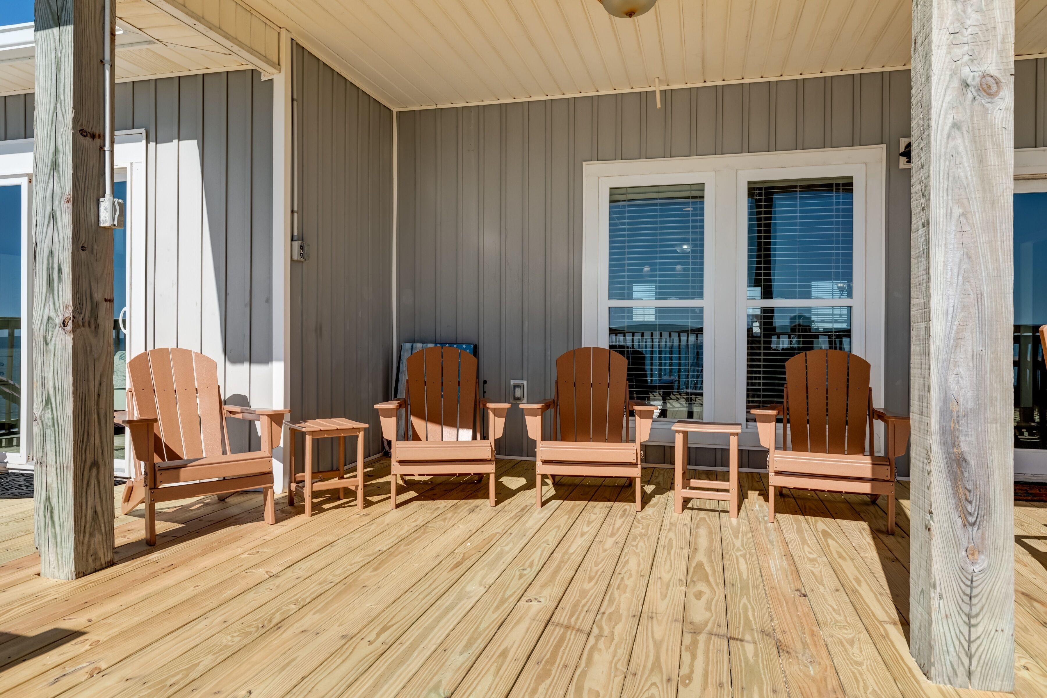 Eine überdachte Terrasse im Freien mit vier Holz-Adirondack-Stühlen und einem kleinen Tisch, Holzsäulen und einem Fenster mit Jalousien.