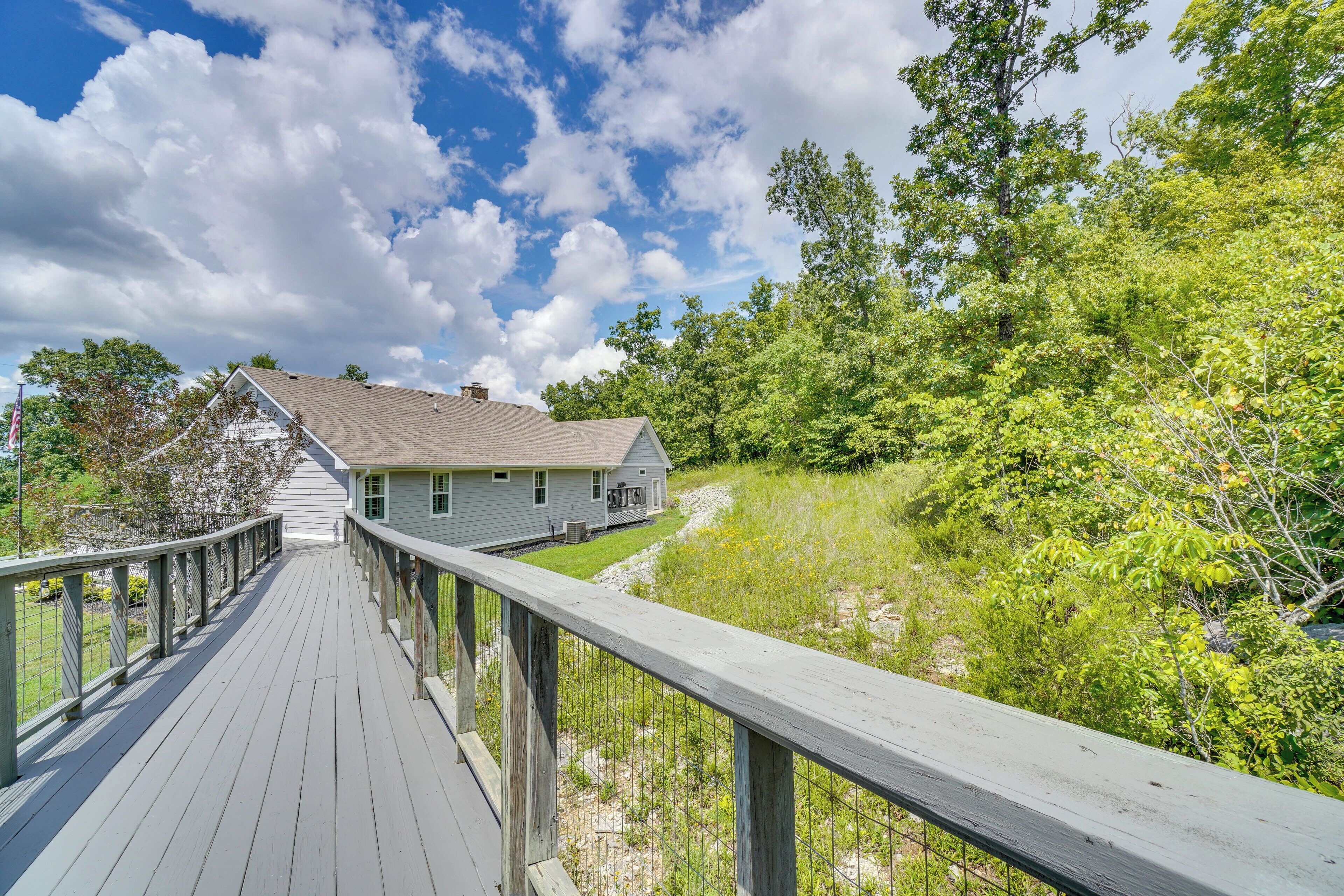 A wooden deck leading to a house with a gray roof, surrounded by lush greenery and trees.