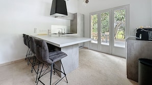 A modern kitchen with a central island, bar stools, a built-in oven, and a view of the outdoor patio through glass doors. - Villa 2 Chambres Avec Jacuzzi (Bonifacio)