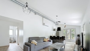 A modern living room with a grey sofa, a coffee table, and a TV. There is a kitchen area with a black stove hood and a dining table with chairs. - Villa 2 Chambres Avec Jacuzzi (Bonifacio)