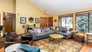 A living room with a sectional sofa, a round coffee table, and a patterned rug. There are framed pictures on the wall and a large window with a view of trees.