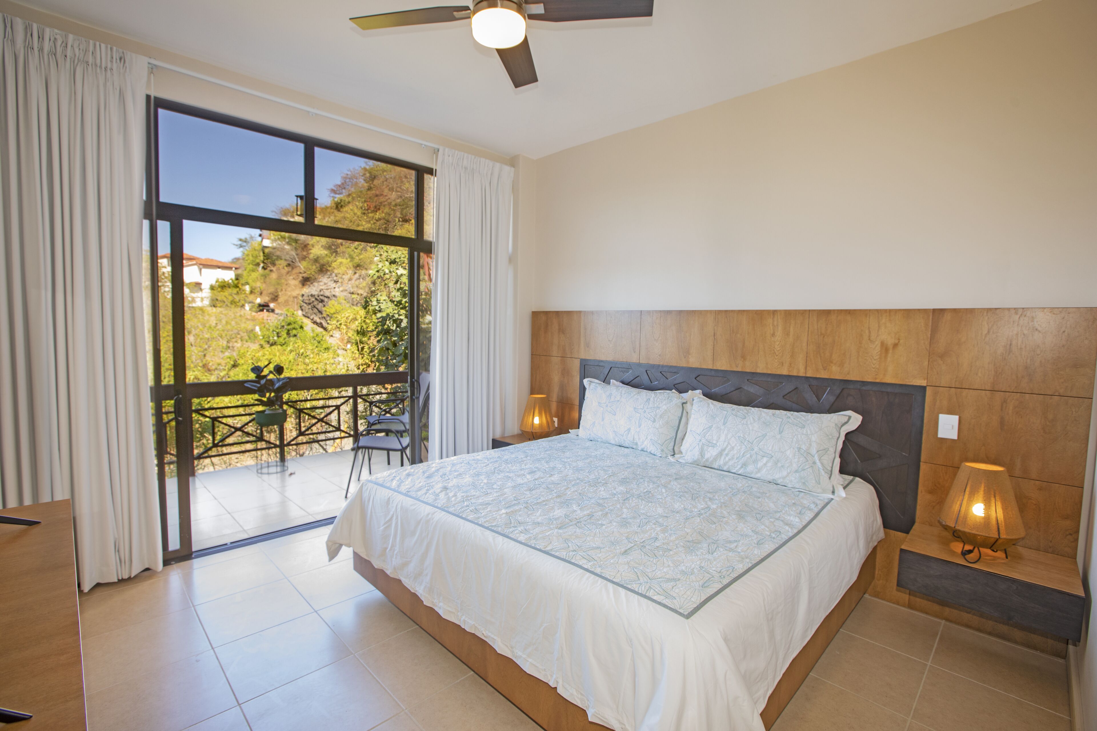 A bedroom with a large bed, a ceiling fan, a nightstand with a lamp, and a view of a balcony with outdoor furniture.
