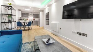 A modern living room with a blue sofa, a coffee table with a remote, a TV mounted on the wall, and a kitchen in the background. - Premium Central Apartment | Free Parking | WiFi (Hampshire)