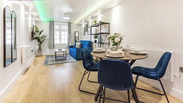 A modern dining area with a round table set for four, blue upholstered chairs, and a blue sofa in the background. - Premium Central Apartment | Free Parking | WiFi (Hampshire)
