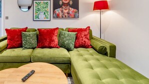A green sofa with red and green cushions, a round wooden coffee table, a wall clock, a framed floral painting, a floor lamp, and a framed abstract art piece. - Groups/Contractors, Pet-friendly, Smart TV, WiFi (Farnborough)