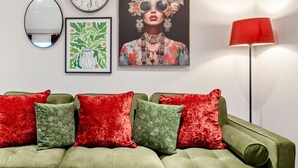 A green sofa with red and green cushions, a wall clock, a framed floral painting, a mirror, and a red floor lamp. - Groups/Contractors, Pet-friendly, Smart TV, WiFi (Farnborough)