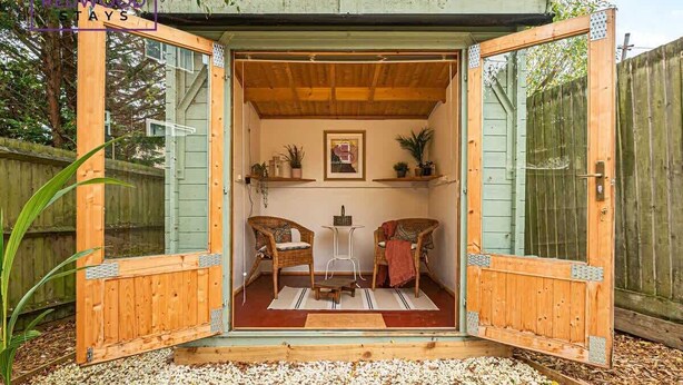 Una cabana de fusta acollidora amb una porta gran oberta, dues cadires de vimetla, una taula petita i un quadre al mur.