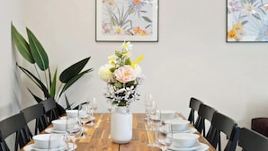 A dining table set for six with white plates, bowls, and wine glasses, surrounded by black chairs. A white vase with flowers is placed in the center. - Huge House, Contractors & Families, Free Parking (Hampshire)